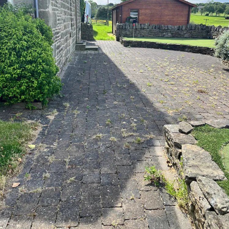 Block paving beside a stone house before pressure washing, with weeds growing through the joints