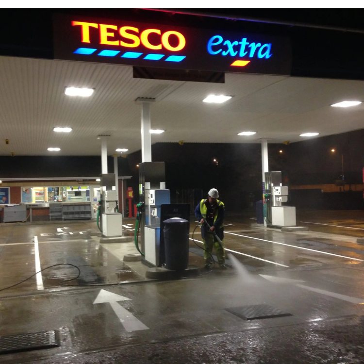Operative pressure washing a petrol station forecourt at night under the canopy lights