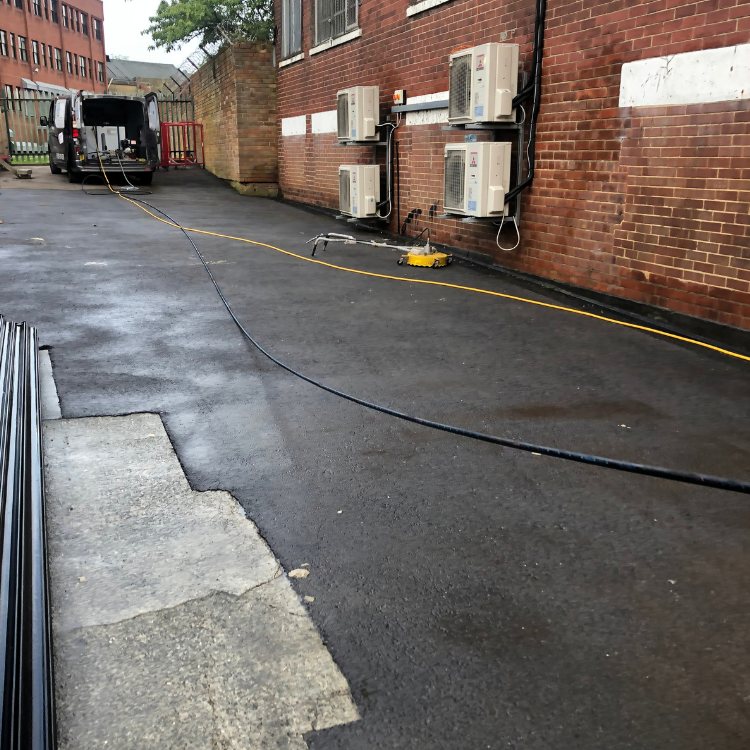 Rotary surface cleaner on wet tarmac beside a brick wall with air conditioning units